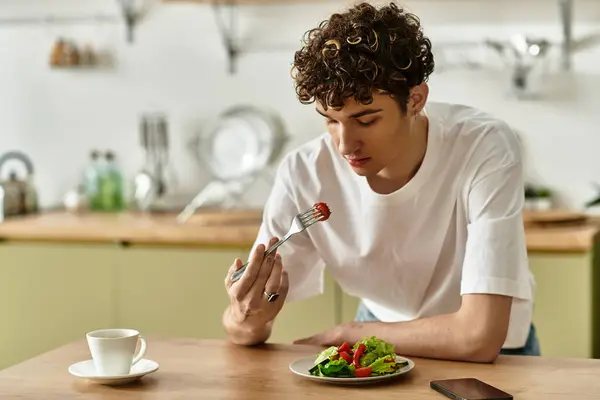 In einer stilvollen Küche genießt ein junger Mann mit lockigem Haar einen bunten Salat, während er Kaffee schlürft. — Stockfoto