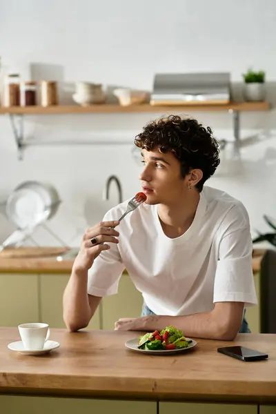 In einer eleganten Küche genießt ein gutaussehender junger Mann einen frischen Salat, verloren in Gedanken, mit einer Gabel in der Hand. — Stockfoto
