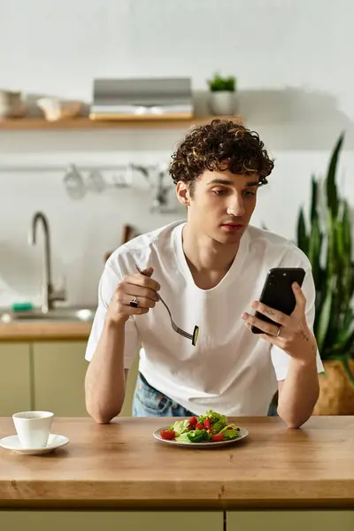In a bright kitchen, a handsome young man checks his phone while savoring a fresh salad and coffee. — Stock Photo