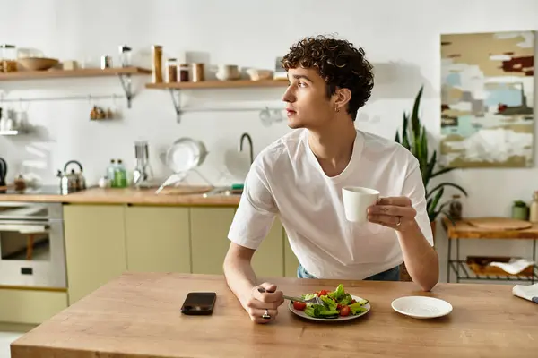 In a stylish kitchen, a handsome young man savors a fresh salad and coffee, gazing thoughtfully out the window. — Stock Photo