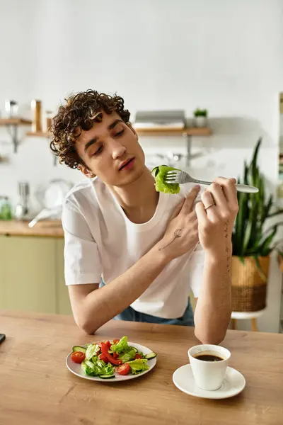 In a stylish kitchen, a handsome young man savors a salad while sipping coffee, embracing healthy living. — Stock Photo