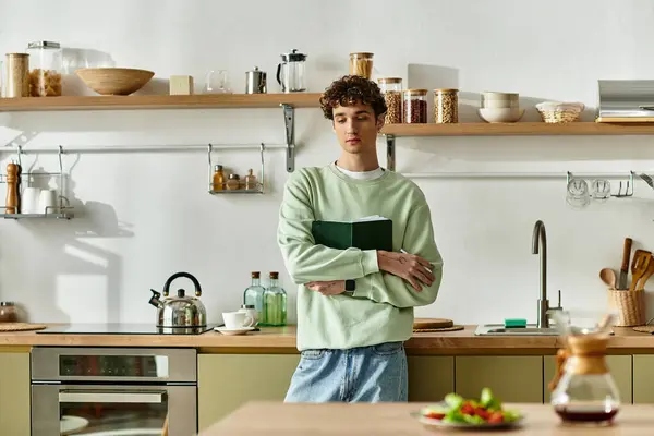 In a contemporary kitchen, a handsome young man with curly hair reflects on his recipe while holding a book. — Stock Photo
