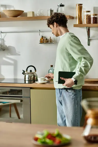 Un jeune homme aux cheveux bouclés verse du café dans une tasse tout en s'habillant élégamment dans une cuisine contemporaine. — Photo de stock