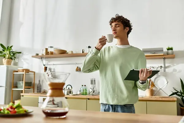 In a bright kitchen, a handsome young man sips coffee, thoughtfully checking his recipe for breakfast. — Stock Photo