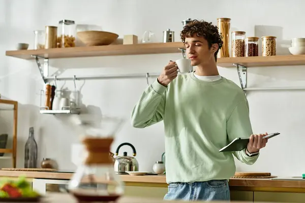 Ein gutaussehender junger Mann mit lockigem Haar schlürft Kaffee und erkundet sein Tablet in einer schicken, modernen Küche. — Stockfoto