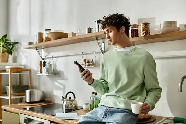 In a stylish kitchen, a handsome young man casually sips his coffee, focused on his smartphone. — Stock Photo