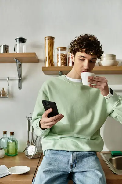 Der gutaussehende junge Mann nippt an seinem Kaffee und benutzt sein Telefon, umgeben vom Charme und Stil einer modernen Küche. — Stockfoto