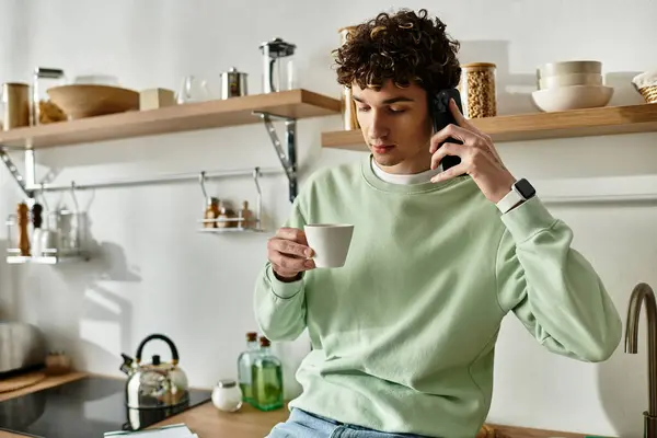 In a stylish kitchen, a handsome young man savors a drink while chatting on the phone, exuding relaxed charm. — Stock Photo