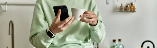 Un bel giovanotto in una cucina elegante tiene una tazza di caffè e guarda il suo smartphone per controllare i messaggi. — Foto stock