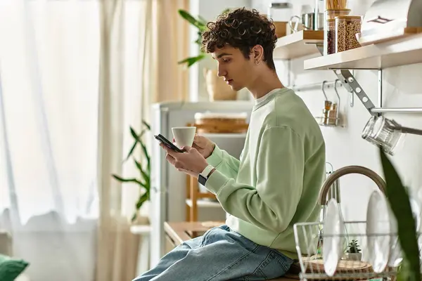 A handsome young man sips coffee, engaged with his phone in a bright, modern kitchen setting. — Stock Photo