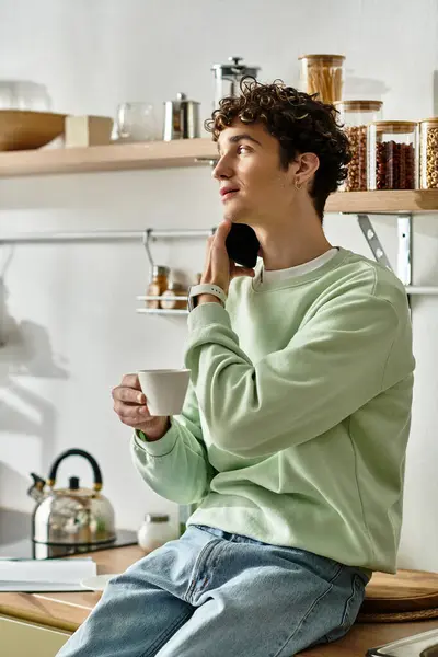A handsome young man relaxes with a cup of coffee, chatting on the phone in a stylish kitchen. — Stock Photo