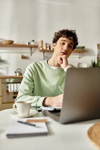 A handsome young man thoughtfully writes in his notebook while focused on his laptop in a contemporary apartment. — Stock Photo