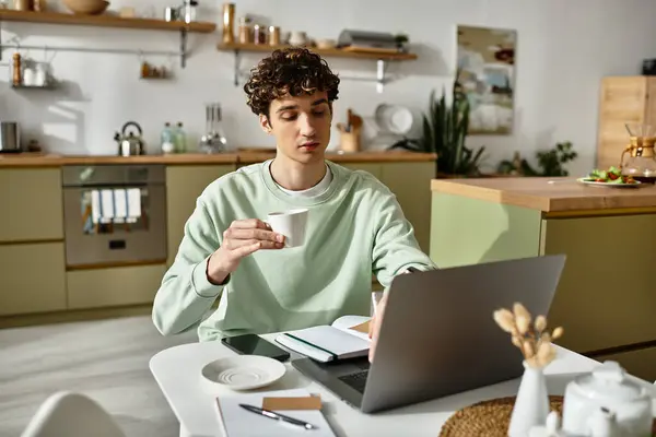 A handsome young man sips coffee from a cup while focused on his laptop in a contemporary home setting. — Stock Photo