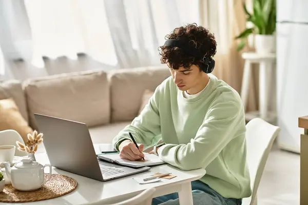 In a bright, modern apartment, a young man with curly hair is intently working on his laptop. — Stock Photo