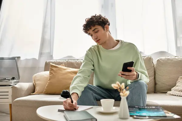 Ein fokussierter junger Mann macht sich Notizen und benutzt sein Handy, während er in einer lichtdurchfluteten modernen Wohnung arbeitet. — Stockfoto