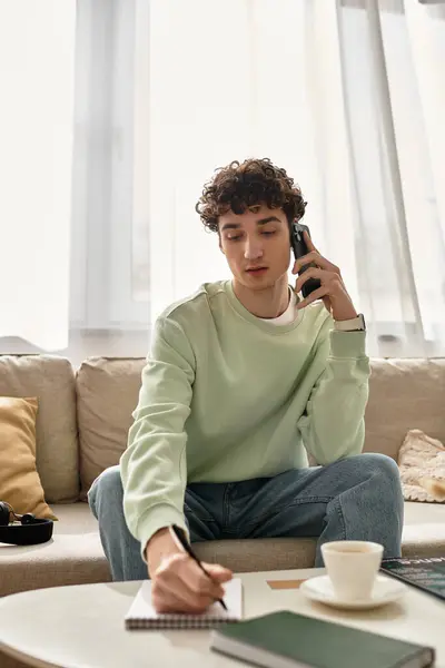 A young man sits comfortably in a stylish apartment, balancing work and a phone call while jotting notes. — Stock Photo