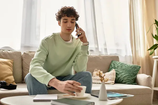 A young man with curly hair is working from home in a stylish apartment while sipping coffee and making a call. — Stock Photo