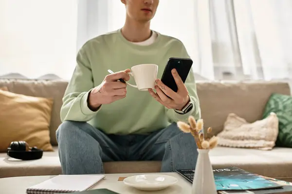 A young man relaxes on his couch, sipping coffee while engaging with his smartphone in a cozy apartment. — Stock Photo