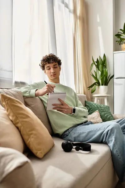 A handsome young man writes in a notepad from his comfortable sofa in a stylish apartment filled with plants. — Stock Photo