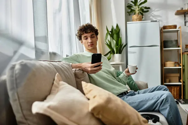 A handsome young man relaxes on his couch, sipping coffee and checking his phone in a stylish living space. — Stock Photo