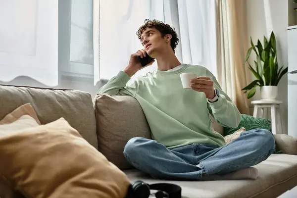 With a cup in hand, a young man with curly hair connects over the phone in his modern, cozy space. — Stock Photo