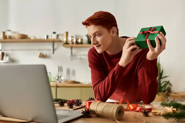 A young man wearing a red sweater excitedly reveals a Christmas gift while sitting at his laptop. — Stock Photo