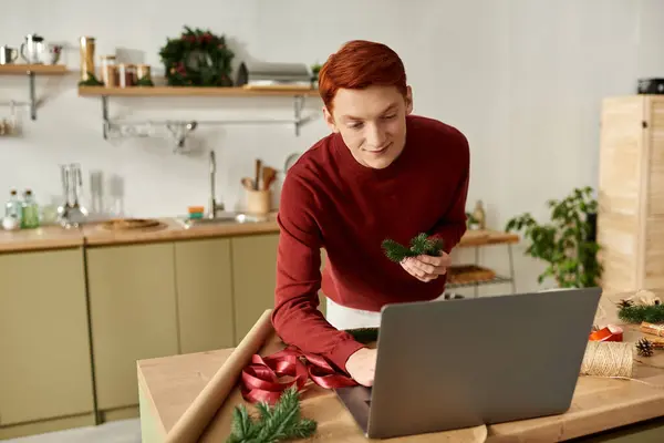 A young man is preparing for Christmas in his kitchen, engaging with his laptop and decorations. — Stock Photo