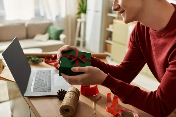 Ein junger Mann lächelt, als er an seinem Schreibtisch, umgeben von festlichem Dekor, ein grünes Weihnachtsgeschenk einwickelt.. — Stockfoto