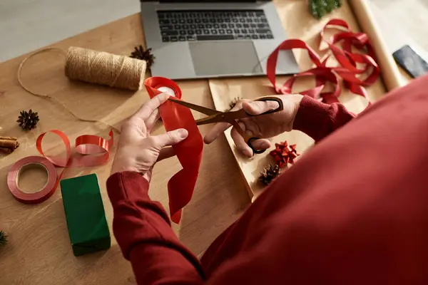 El joven corta cuidadosamente la cinta roja para preparar sus regalos de Navidad en un espacio de trabajo festivo.. - foto de stock