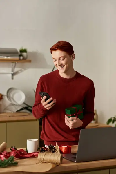 Ein fröhlicher junger Mann mit einem Geschenk lächelt, als er sein Telefon in einer gemütlichen Küche checkt. — Stockfoto