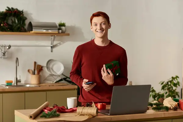 A young man with red hair is smiling as he holds a gift while surrounded by Christmas decorations. — Stock Photo