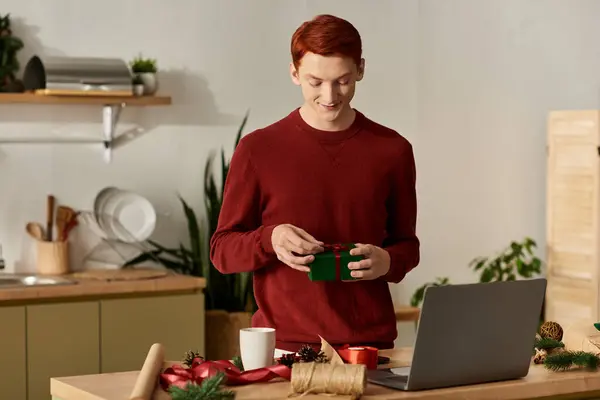A young man in a red sweater excitedly unwraps a gift while preparing for Christmas in his kitchen. — Stock Photo