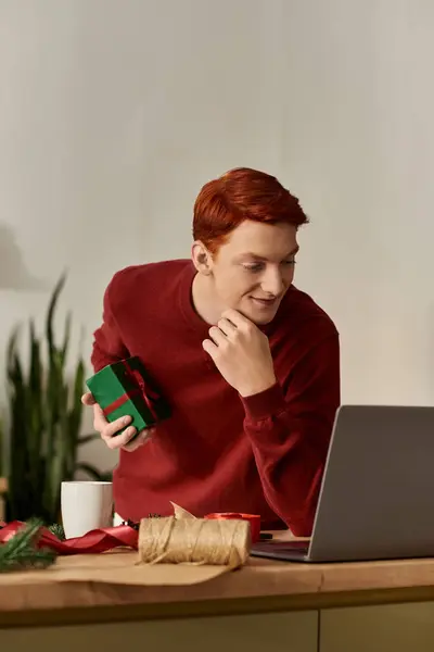 A cheerful young man in a cozy setting is wrapping a Christmas gift while using his laptop. — Stock Photo