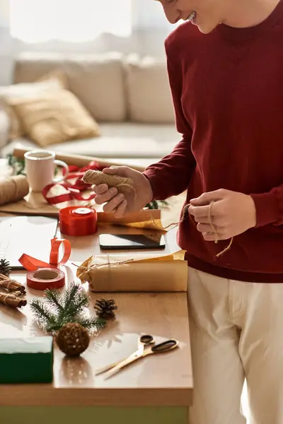 Um jovem gosta de embrulhar presentes de Natal em uma mesa festiva cheia de decorações e suprimentos. — Fotografia de Stock