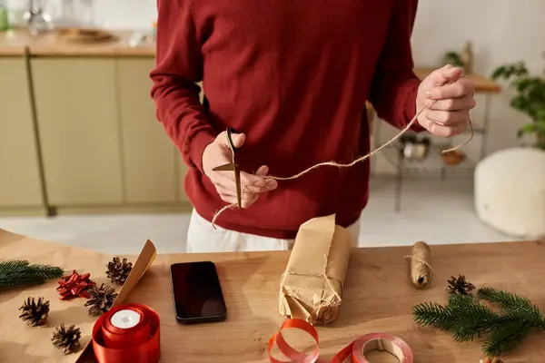 El joven ata cuidadosamente el cordel alrededor de un regalo de Navidad bellamente envuelto en casa. - foto de stock