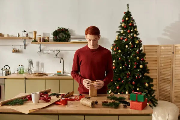 In a warm kitchen, a young man carefully wraps gifts while surrounded by holiday decorations. — Stock Photo