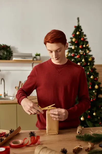 Un giovane uomo con un maglione rosso avvolge un regalo di Natale con attenzione e cura in una stanza accogliente. — Foto stock