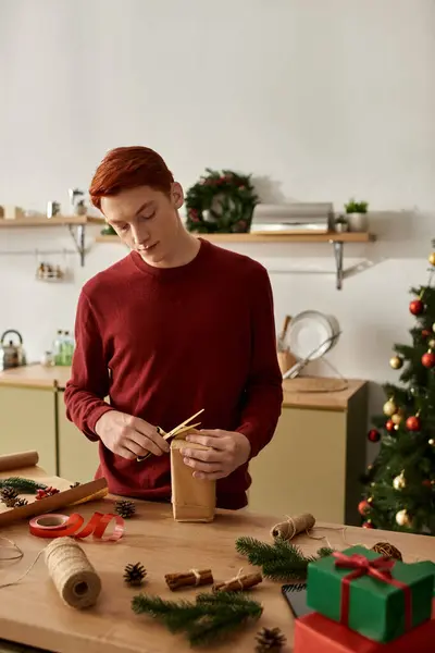 Il giovane, vestito con abiti festivi, si concentra sull'avvolgere un regalo con cura durante le vacanze.. — Foto stock