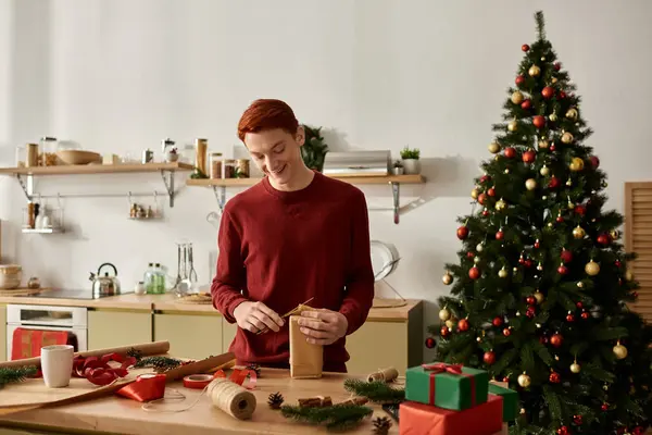 In a cheerful kitchen, a young man wraps holiday gifts while surrounded by Christmas decor. — Stock Photo