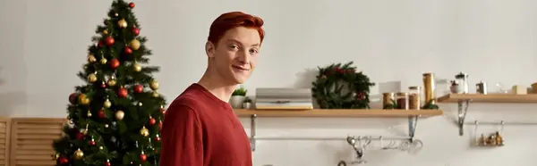 Um jovem alegre fica perto de uma árvore de Natal decorada em um ambiente de cozinha quente. — Fotografia de Stock