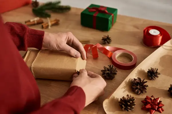 Un joven envuelve cuidadosamente los regalos para Navidad rodeado de decoraciones y suministros navideños.. - foto de stock