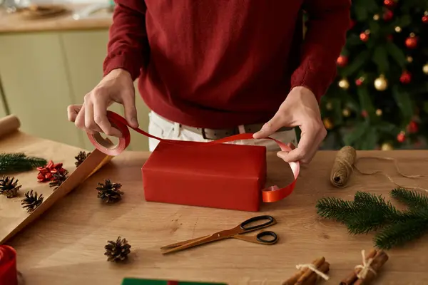 Un joven envuelve cuidadosamente un regalo con cinta en medio de decoraciones navideñas y conos de pino. - foto de stock