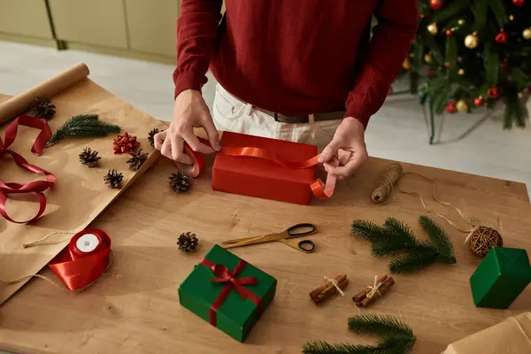 Un joven envuelve meticulosamente los regalos de Navidad en una mesa adornada con decoraciones festivas. - foto de stock