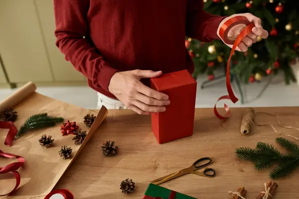 Le jeune homme enveloppe soigneusement un cadeau rouge vibrant entouré de décorations et de fournitures de vacances. — Photo de stock