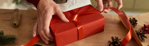 El joven está cuidadosamente envolviendo un brillante regalo y agregando una cinta encantadora para Navidad.. - foto de stock