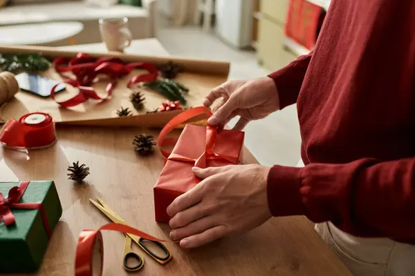 El joven envuelve cuidadosamente un regalo rojo de Navidad rodeado de decoraciones y suministros festivos.. - foto de stock