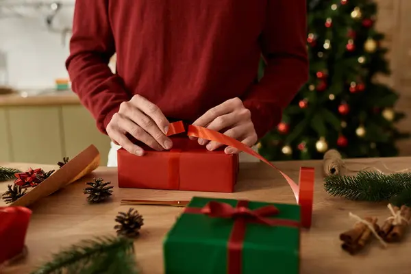 A young man is carefully wrapping a Christmas gift surrounded by holiday decorations and greenery. — Stock Photo