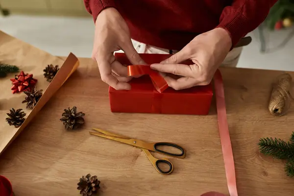 Un joven está atando una hermosa cinta alrededor de un regalo rojo cuidadosamente envuelto en un ambiente festivo. - foto de stock