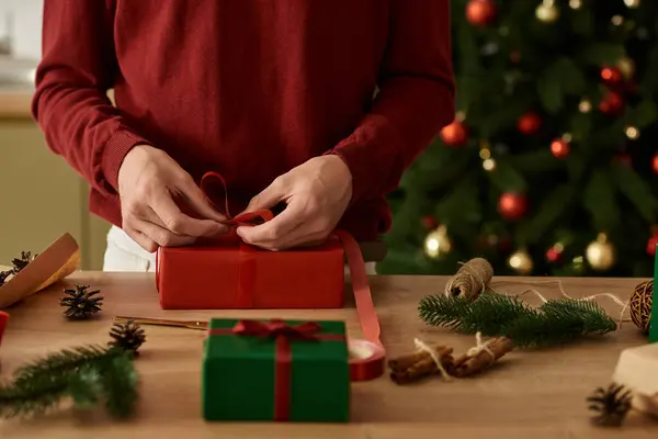 El joven envuelve hábilmente los regalos en una mesa de madera mientras está rodeado de una decoración festiva.. - foto de stock