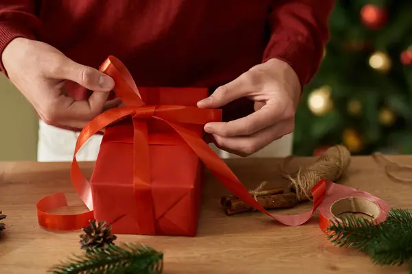 A young man is tying a vibrant ribbon around a beautifully wrapped Christmas gift while smiling. — Stock Photo